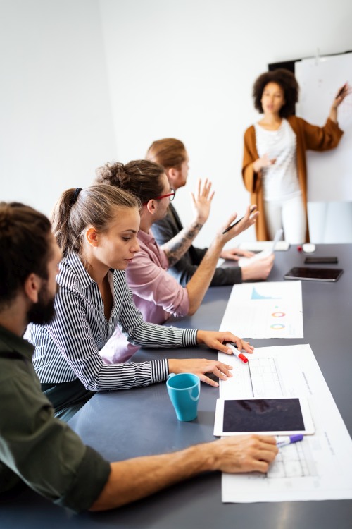 Business people working with HR tools at a meeting. 4 people sit at large table and a woman points to HR analytics on a board.